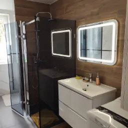 Nowoczesna łazienka z kabiną prysznicową typu walk-in, umywalką z szafką, podświetlanymi lustrami i płytkami imitującymi drewno na ścianie, uzupełniona o pralkę.