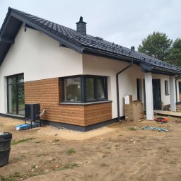 USŁUGI REMONTOWE MARCIN WIECZOREK - Nowoczesny dom jednorodzinny z białą elewacją i ciemnym dachem, częściowo wykończony drewnianą okładziną. Widoczne czarne ramy okienne i teren budowy.