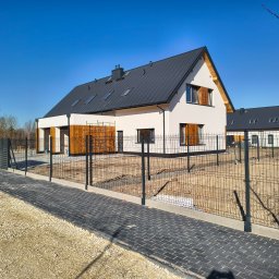 USŁUGI REMONTOWE MARCIN WIECZOREK - Nowoczesny dom jednorodzinny z białą elewacją, ciemnoszarym dachem z lukarnami i drewnianymi akcentami. Przed domem panelowe ogrodzenie i ułożona kostka brukowa.