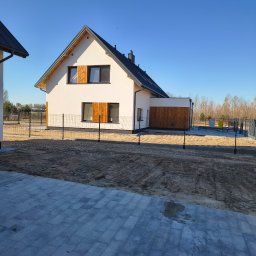 USŁUGI REMONTOWE MARCIN WIECZOREK - Nowoczesny dom z białą elewacją, ciemnym dachem, drewnianymi okiennicami i panelami, otoczony siatkowym ogrodzeniem, z częściowo wybrukowanym podjazdem i niezagospodarowanym terenem.