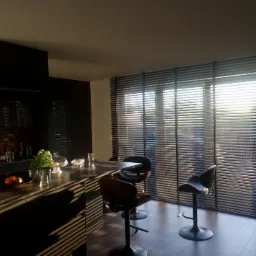 Ciemne poziome żaluzje w nowoczesnej kuchni z wyspą i wysokimi stołkami barowymi, oświetlone popołudniowym słońcem wpadającym przez okno.