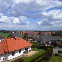 Panorama osiedla domów jednorodzinnych z czerwonymi i szarymi dachami pod błękitnym niebem z obłokami. Widok z góry na zadbane ogrody i pola w tle.