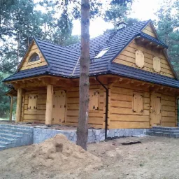 Dwukondygnacyjny dom z bali z ciemną dachówką i kamiennymi schodami wejściowymi, otoczony drzewami. Widoczny fragment placu budowy.