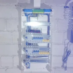 Rozdzielnica elektryczna z zainstalowanymi bezpiecznikami i przewodami, zamontowana na ścianie z białej cegły. Widoczny również moduł TMS Duo P z przewodami.