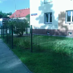 Nowoczesne ogrodzenie panelowe w kolorze zielonym, otaczające posesję z budynkiem mieszkalnym i zielonym trawnikiem. Widoczny fragment chodnika i ulicy.