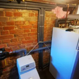 Piwnica z instalacją centralnego ogrzewania: nowy piec CO, rury miedziane z izolacją, zmiękczacz wody, ceglana ściana z fragmentem ocieplenia.