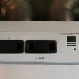 Centrala wentylacyjna Vaillant recoVAIR z widocznymi filtrami i panelem sterowania, zamontowana pod sufitem z widocznymi kanałami wentylacyjnymi.