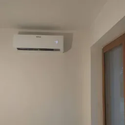 Biały klimatyzator marki Vaillant zamontowany na jasnej ścianie w pomieszczeniu, widoczny wyświetlacz z temperaturą 16 stopni Celsjusza, obok okno z drewnianą ramą.