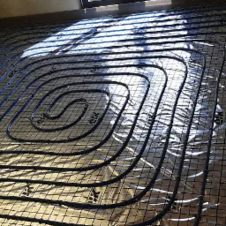 Niebieskie rury ogrzewania podłogowego ułożone na srebrnej folii izolacyjnej, zamocowane do czarnej siatki na tle surowego betonu i okna z refleksami światła.