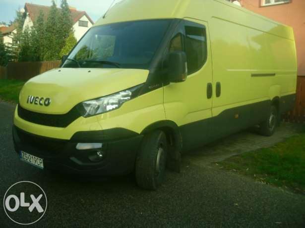 Żółto-szary bus Iveco z widoczną tablicą rejestracyjną, zaparkowany na ulicy. Widok z boku, z przodu. Tło stanowią domy i drzewa.