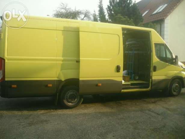 Żółty bus z otwartymi drzwiami bocznymi, wewnątrz widoczny wózek transportowy. Dwukolorowa karoseria, ciemnoszary dół, jasny żółty góra. Trawa i budynek w tle.