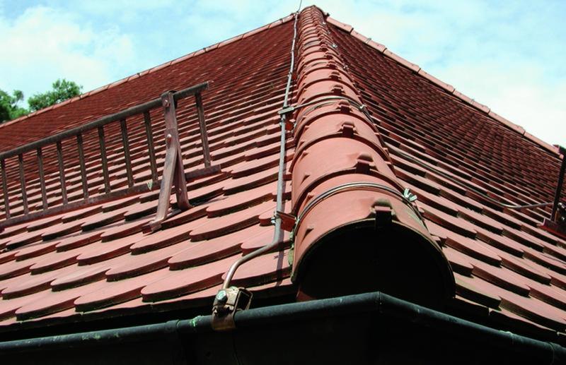 Czerwony dach pokryty dachówką z zamontowanym systemem odgromowym, widoczne metalowe elementy i przewody oraz drabinka kominiarska.