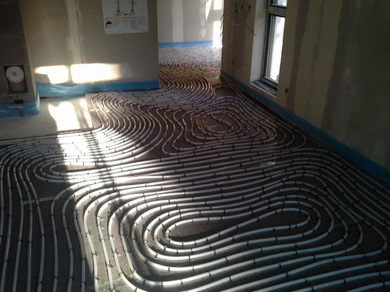 Białe rury ogrzewania podłogowego ułożone spiralnie na izolacji w surowym wnętrzu, przygotowane do zalania wylewką. Widoczne okno i fragmenty ścian.