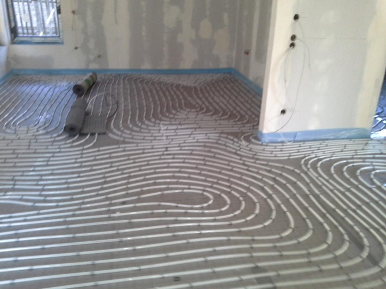 Białe rury ogrzewania podłogowego ułożone w pętle na szarym podłożu, widoczne rolki izolacji i nieotynkowane ściany z niebieską folią ochronną.