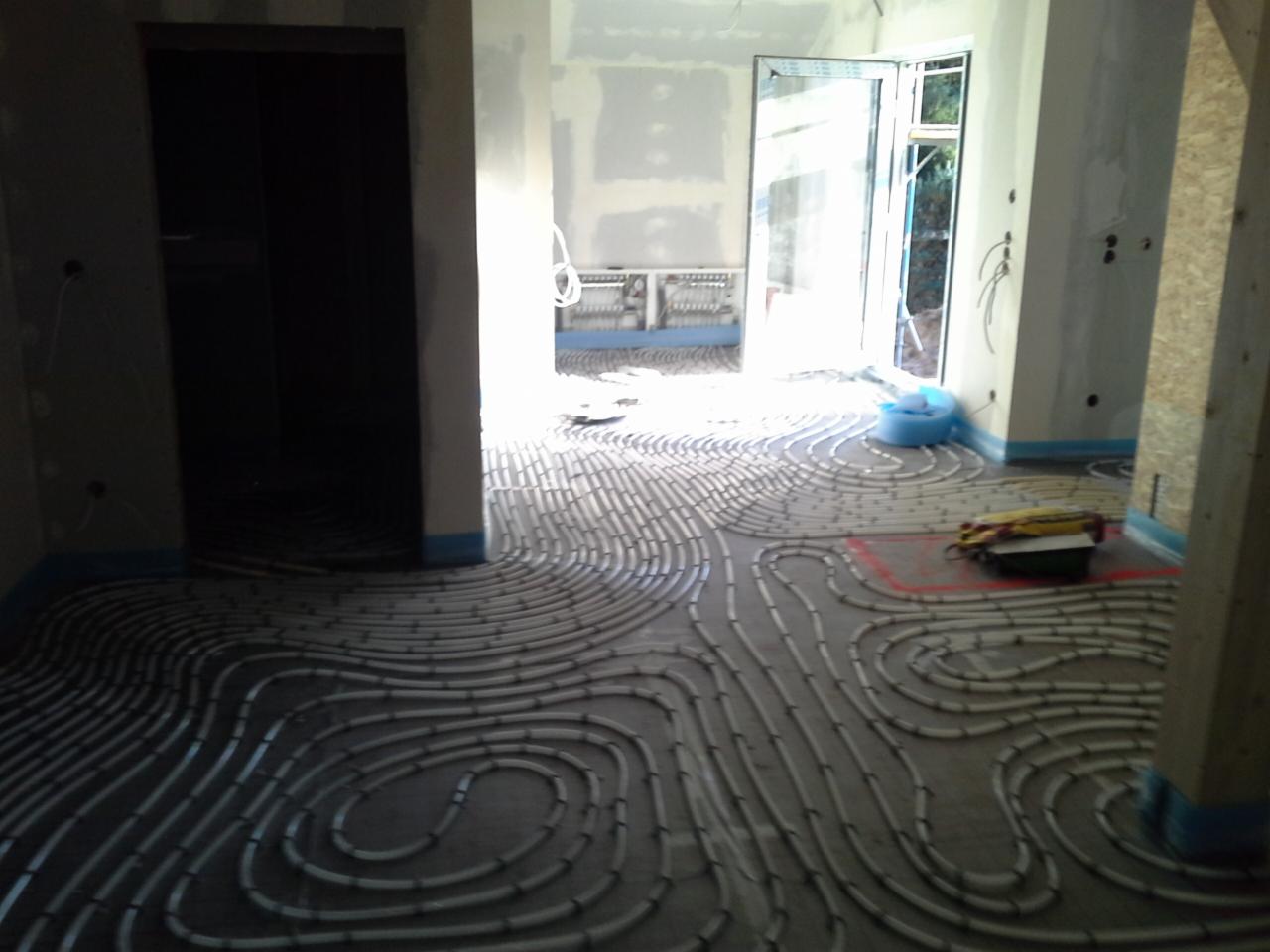 Rozłożone rury ogrzewania podłogowego w surowym wnętrzu, widoczne narzędzia i materiały, przygotowanie do wylewki, błękitna taśma zabezpieczająca.