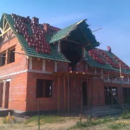 Budowa domu z czerwonej cegły: częściowo położony dach z czerwonej dachówki, widoczny murarz na dachu, drewniane rusztowania i niezadaszone okna.