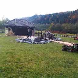 Ogród z altaną o kamiennej elewacji i ciemnym dachu, ozdobnym mostkiem nad fontanną, placem zabaw i zielonym trawnikiem na tle zalesionego wzgórza.