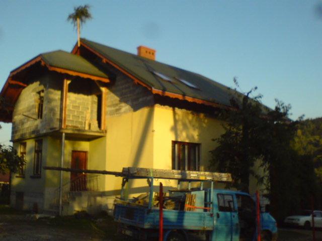 Dom w trakcie budowy z częściowo wykończoną elewacją i dachem. Na dachu widoczne gniazdo. Na pierwszym planie samochód dostawczy z drabiną.