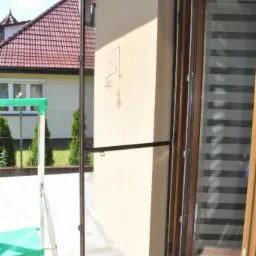 Otwarta moskitiera ramkowa w Zawadzie, zamontowana przy drzwiach balkonowych. Widoczne sandały i drabina. W tle dom z czerwonym dachem i zielenią.