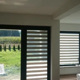 Nowoczesne okno z zamontowanymi roletami w biało-beżowe pasy, widok na ogród z tujami i pole za oknem, surowa żarówka zwisająca z sufitu.