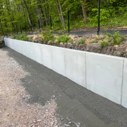 Wzniesiony betonowy mur oporowy z żółtą koparką JCB w tle, widoczne świeżo wyrównane podłoże z kamienia i ziemi.