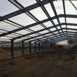 Wnętrze w trakcie budowy: stalowa konstrukcja hali z kolumnami i dachem kratownicowym. W tle widoczna maszyna budowlana i materiały.