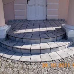 Wykończone, półokrągłe schody wejściowe z szarych płytek ceramicznych z ciemną fugą, prowadzące do białych drzwi wejściowych z kolumnami po bokach, na tle różowej elewacji.