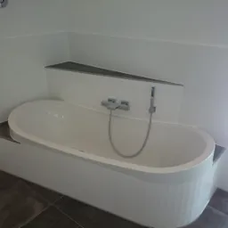 Nowoczesna wanna owalna obudowana białymi płytkami, z szarą półką i armaturą, na podłodze ciemne płytki.