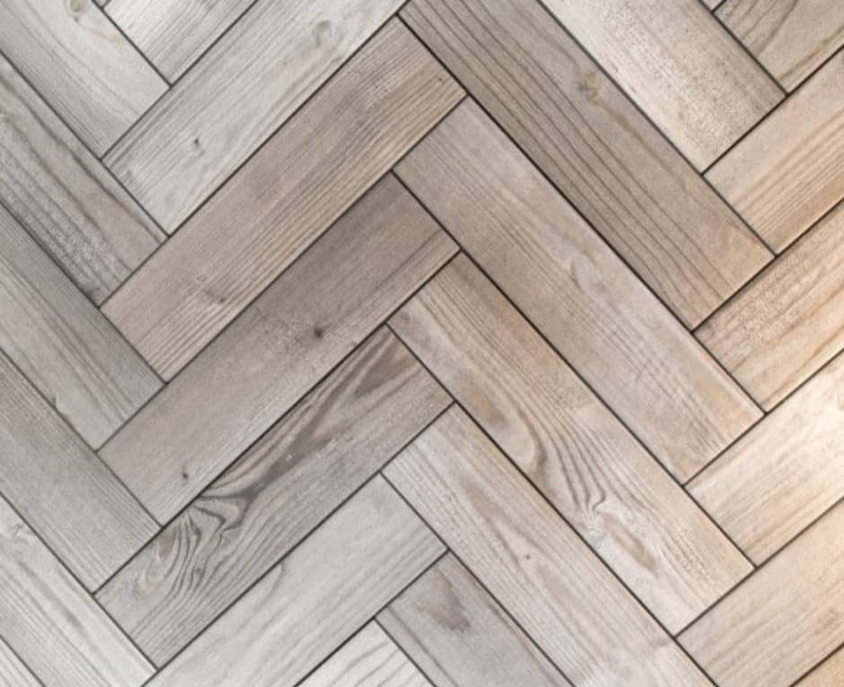 Jasna, drewnopodobna podłoga ułożona we wzór jodełki francuskiej, z widoczną strukturą drewna i ciemniejszymi fugami, oświetlona z prawej strony.