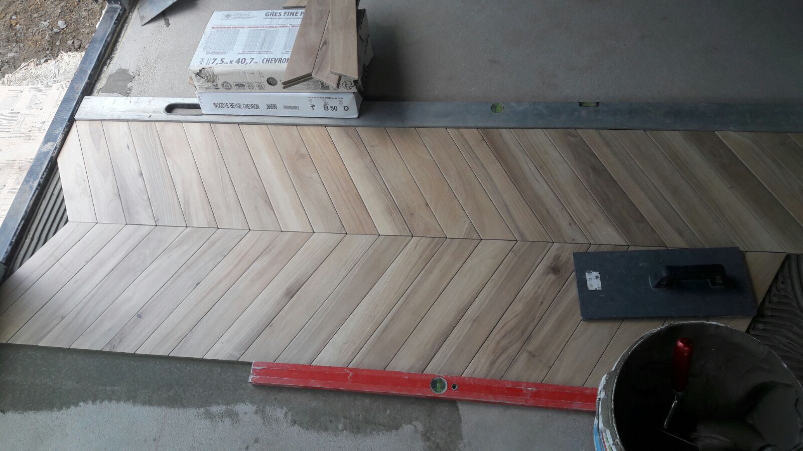 Ułożone płytki gresowe w jodełkę na podłodze, widoczny poziomica, paca do rozprowadzania kleju i wiadro z zaprawą, pudełko z płytkami w tle.