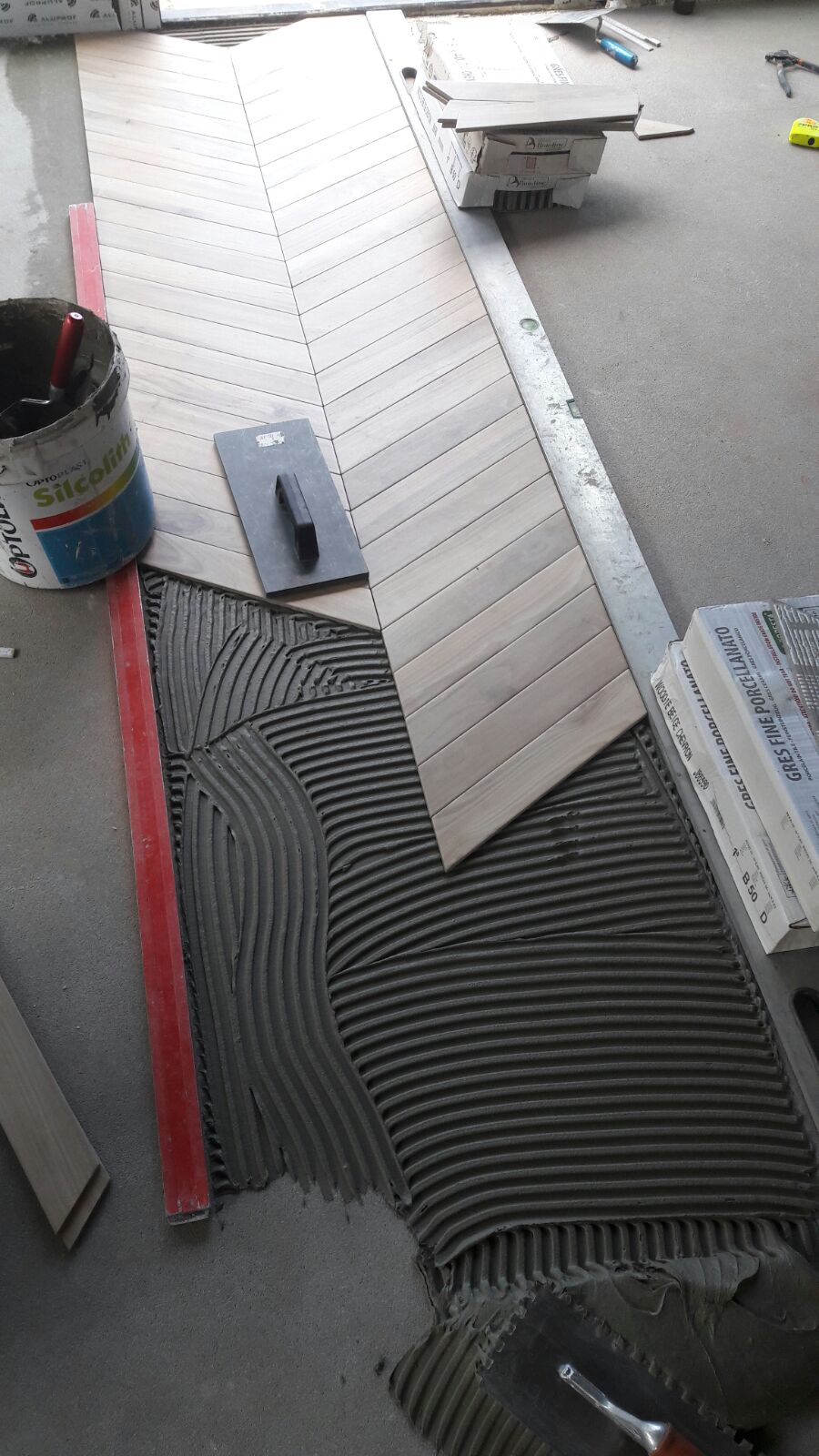 Układanie podłogi z płytek drewnopodobnych w jodełkę; widoczny fragment ułożonej podłogi, paczki płytek Gres Fine Porcellanato, wiadro kleju Silcolith, poziomica, szpachelka i rozprowadzony klej...