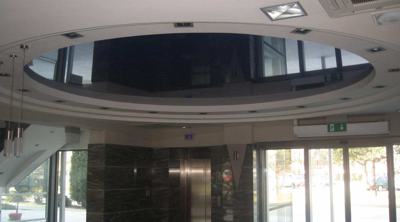 Błyszczący, czarny sufit w kształcie okręgu, odbijający wnętrze biura, z punktowymi lampami i dekoracyjnymi listwami, widok z dołu w holu biurowca z windą i szklanymi drzwiami.