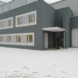 Szary budynek po tynkowaniu w zimowej scenerii. Białe ramy okienne i brama garażowa kontrastują ze śniegiem. Drabina przy wejściu sugeruje prace wykończeniowe.