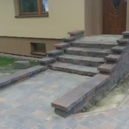 Schody wejściowe i chodnik z kostki w odcieniach szarości i brązu, z widocznymi niedociągnięciami w postaci ziemi przy obrzeżach.