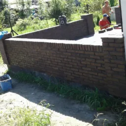 Wznoszenie muru z cegły klinkierowej, widoczni dwaj mężczyźni podczas pracy, jeden w czerwonej koszulce układa cegły, drugi bez koszulki stoi obok, na ziemi wiadro i narzędzia, w tle zieleń drzew...