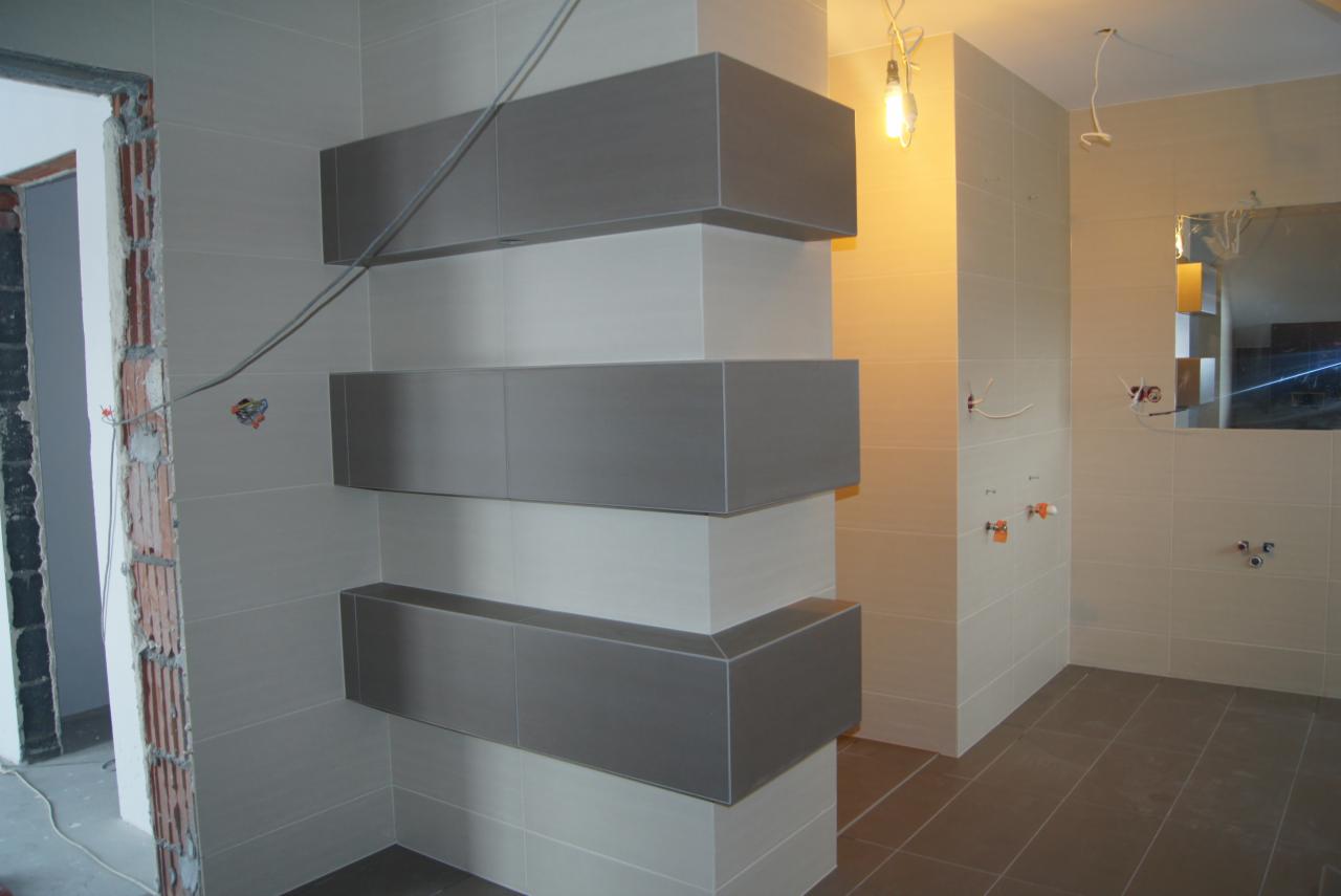 Łazienka w trakcie wykańczania: szare, prostokątne szafki na ścianie z jasnych płytek, widoczne instalacje, surowy oścież drzwi i ciemna podłoga.