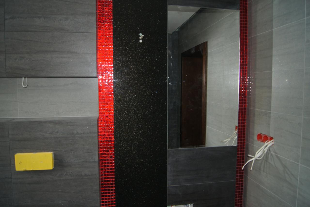 Wykończona łazienka z szarymi płytkami imitującymi drewno, czarnym panelem i pionowym pasem czerwonej mozaiki. Widoczne gniazdka elektryczne i lustro.