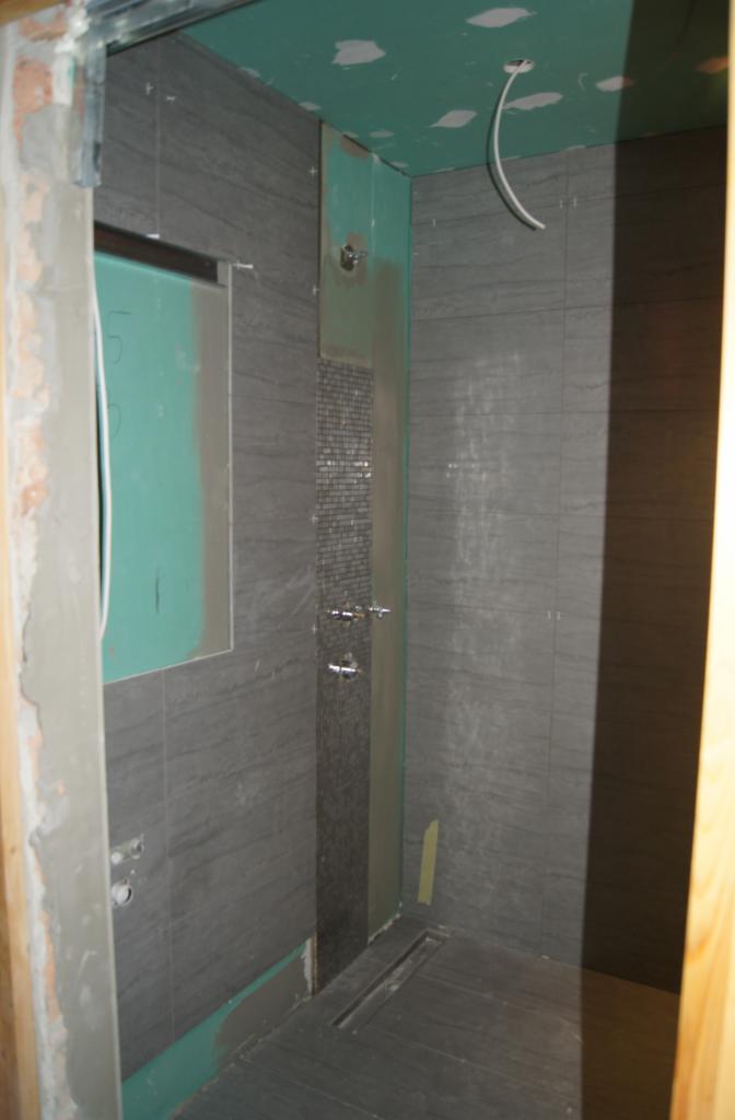 Szara łazienka w trakcie remontu: płytki imitujące kamień, mozaika pod prysznicem, widoczne prace wykończeniowe, surowy stan, odpływ liniowy w podłodze.