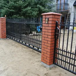 Czarna metalowa brama wjazdowa z elementami dekoracyjnymi, osadzona na ceglanych słupkach, z widoczną skrzynką na listy na jednym ze słupków, na tle domu z balkonem i drabiny opartą o ścianę.