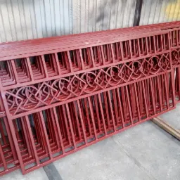 Stos pomalowanych na czerwono, metalowych elementów ogrodzenia z ozdobnymi, okrągłymi wzorami, leżących na betonowej podłodze w warsztacie.