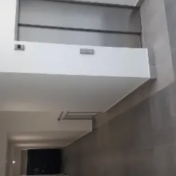 Wnętrze korytarza z szarymi płytkami na podłodze i ścianach, białe tynki na suficie i górnej części ściany, oświetlenie punktowe i metalowy wieszak na ręczniki.