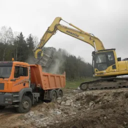 Żółta koparka Komatsu PC 240 NLC załadowuje urobek do pomarańczowej wywrotki MAN na placu budowy, widoczne drzewa w tle.