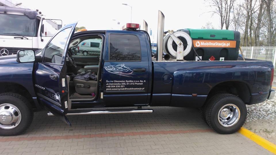 Niebieski pickup z otwartymi drzwiami, wyposażony w mobilną stację paliw z zielonym zbiornikiem i wężem na pace, zaparkowany na brukowanym placu.