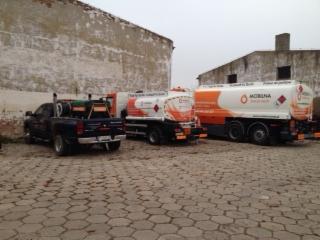 Trzy pojazdy: czarny pickup z zabudową oraz dwie cysterny z pomarańczowymi akcentami i logo, zaparkowane na brukowanym placu przy budynku.