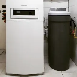 Biały piec grzewczy Buderus z wyświetlaczem temperatury i szary zmiękczacz wody Trinity Home w pomieszczeniu z białymi kafelkami.