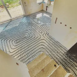 Widok z góry na rozłożone rury grzewcze na siatce w pomieszczeniu z dużym oknem i surowymi ścianami, przygotowane do zalania wylewką.