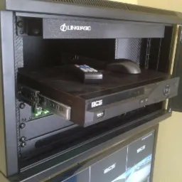 Serwerownia z rejestratorem BCS na wysuwanej półce, pilot i mysz komputerowa na górze, widoczne okablowanie i monitor poniżej.