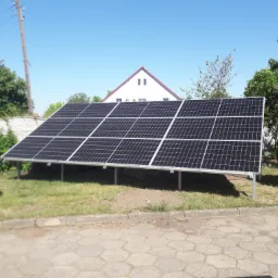 Instalacja fotowoltaiczna na trawniku przed domem z czerwoną dachówką, widok z poziomu chodnika.