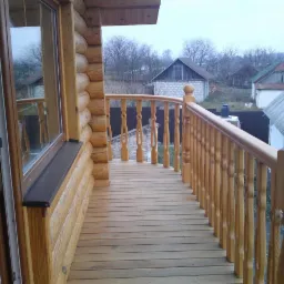 Drewniany balkon z balustradą w domu z bali, widok z wnętrza, drewniana podłoga, za balustradą widoczne budynki gospodarcze w oddali.