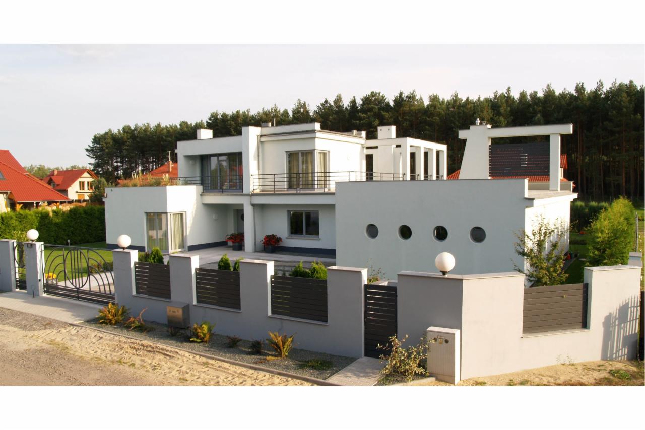 Nowoczesny dom z płotem panelowym i ozdobną bramą. Geometryczna architektura, minimalistyczny design, szara kolorystyka. Widok z zewnątrz na tle zieleni.